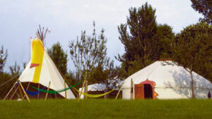 Large 21 ft diameter Hearthworks Tipi with Yellow and Red trim and an Extra Large 24 ft diameter Hearthworks Yurt with bespoke door design set up at a Wedding event with hammocks outside