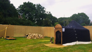 Hearthworks Yurt Sauna with carved wooden door and black canvas cover in enclosed area at festival with screen walls and outdoor shower